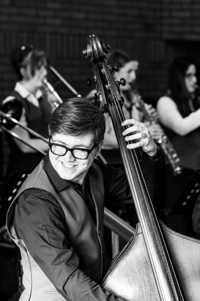 A smiling double bass player mid-rehearsal, with saxophones and trombone behind